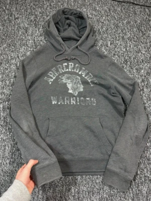  Vintage Abercrombie & fitch hoodie (extremt sällsynt) - Vintage Mörkgrå hoodie från Abercrombie med trycket 'Abercrombie Warriors' och ett motiv på bröstet. Klassisk känguruficka framtill och justerbar huva med dragsko. Mjuk bomullsblandning som känns skön mot huden. Perfekt för en avslappnad streetwear-look.