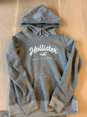 Grå hoodie från Hollister - Storlek XS men skulle säga att den passar folk som har S, köpt för 600. Snygg grå hoodie från Hollister med vit logga och texten 'California' på bröstet. Klassisk känguruficka framtill, ribbade muddar och dragsko i huvan. Perfekt för chill dagar och enkel att matcha med jeans eller mjukisbyxor.