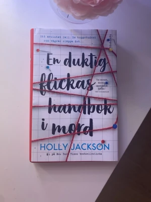 En duktig flickas handbok i mord - En spännande deckare av Holly Jackson med hård pärm i vitt och rött, dekorerad med röda trådar och nålar på omslaget. Boken är på svenska och handlar om en ung tjej som undersöker ett gammalt mordfall. Perfekt för dig som gillar mysterier och thrillers.
