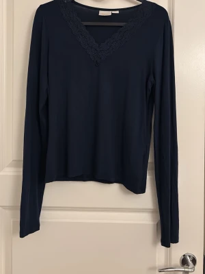 Mörkblå långärmad topp med spets - Mörkblå långärmad topp med v-ringning och dekorativ spetskant runt halsen. Toppens material är mjukt och stretchigt, vilket gör den bekväm att bära. Enkel och stilren design som passar till många olika outfits.