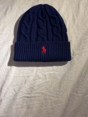 Mörkblå kabelstickad mössa Polo Ralph Lauren - Snygg mörkblå kabelstickad mössa från Polo Ralph Lauren med röd broderad logga framtill. Mössan har uppvikt kant och är gjord i ett mjukt, värmande material. Perfekt för kalla dagar och en stilren look.