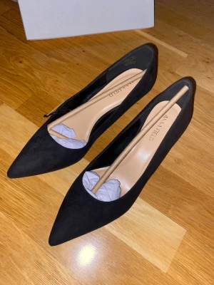 Svarta pumps från Anna Field - Stilrena svarta pumps från Anna Field med spetsig tå och låg klack. Skorna är tillverkade i mocka och har en klassisk, elegant design som passar perfekt till många olika outfits.
