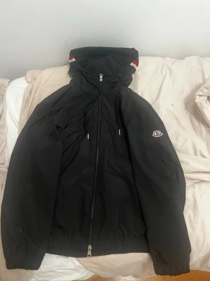 Moncler vindjacka - Säljer en svart vindjacka från Moncler med huva och dragkedja framtill. Jackan har elastiska muddar, broderad Moncler-logga på ärmen och diskreta röda och vita detaljer på huvan. Perfekt för dig som vill ha en stilren och funktionell jacka.
