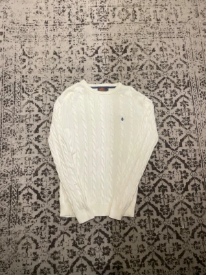 Vit kabelstickad tröja från Morris - Kabelstickad vit tröja från Morris med rund halsringning och diskret blå broderad logga på bröstet. Tröjan har långa ärmar och ribbade muddar. Tillverkad i mjuk bomull för en klassisk och stilren look.