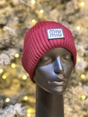 Vinröd ribbstickad mössa från Miu Miu - Snygg vinröd ribbstickad mössa från Miu Miu med en tydlig logotyp framtill. Mössan har en klassisk passform och är tillverkad i ett mjukt, värmande material som passar perfekt för kyliga dagar.