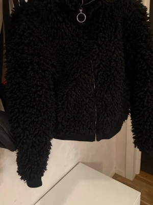 Svart teddyjacka med dragkedja - Mysig svart teddyjacka med fluffig struktur och hög krage. Jackan har en hel dragkedja framtill med en rund metallring som detalj. Ribbstickade muddar vid ärmslut och nederkant ger en skön passform. Perfekt för kyliga dagar. Size är xs men passar till M.