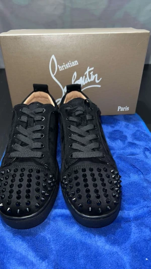 Christian Louboutin svarta sneakers med nitar - Svarta sneakers från Christian Louboutin med coola svarta nitar på tån och klassisk röd sula. Skorna har snörning och är tillverkade i mocka med detaljer i skinn. Perfekt för dig som vill sticka ut med en lyxig och edgy stil