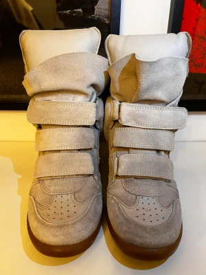 Beige höga sneakers i mocka - Unika beige sneakers med högt skaft och flera kardborreband. Tillverkade i mjuk mocka med rund tå och platt sula i brunt gummi. Skorna har en futuristisk design med extra vaddering runt ankeln och tungan.