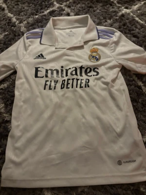 Vit Real Madrid matchtröja Adidas - Säljer en vit Real Madrid matchtröja från Adidas med svart text och klubbmärke på bröstet. Tröjan har krage, lila detaljer på axlarna och är tillverkad i lätt polyester med AEROREADY-teknologi. Perfekt för fotbollsträning eller att bära som supporter.
