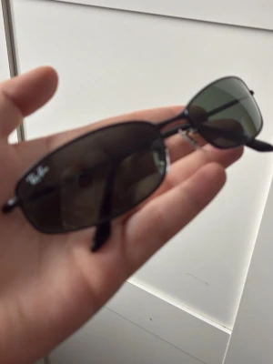 Ray-Ban smala svarta solglasögon - Snygga solglasögon från Ray-Ban med smal, rektangulär form och svarta metallbågar. Glasen är mörktonade och ger en cool retrovibe. Perfekta för dig som vill ha en stilren accessoar med klassisk Ray-Ban-logo på linsen.