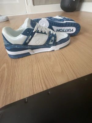 Louis Vuitton blåvita sneakers - Aldrig använda!!! Säljer ett par Louis Vuitton sneakers i blått och vitt med tydliga LV-loggor och monogramdetaljer. Skorna har snörning, platt sula och är tillverkade i en mix av skinn och textil. Snygg kontrast mellan blå denim och vita partier, perfekt för dig som gillar exklusiva sneakers.