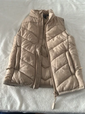 Beige dunväst med dragkedja - Stilren beige dunväst med hög krage och dragkedja framtill. Västen har quiltat mönster och justerbara snören i sidorna för en snygg passform. Perfekt att slänga över en hoodie eller tröja för extra värme. Materialet är lätt och smidigt.