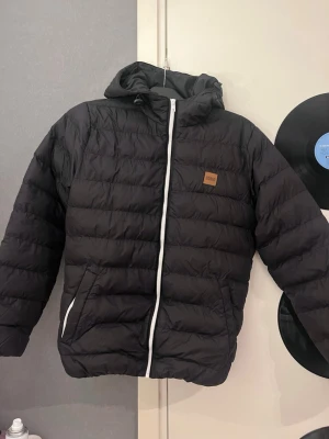 Svart dunjacka med huva från urban classics - Säljer en svart dunjacka från Wear med huva och vit dragkedja. Jackan har quiltad design, två sidofickor och en liten logopatch på bröstet. Perfekt för kalla dagar och enkel att matcha med olika outfits.