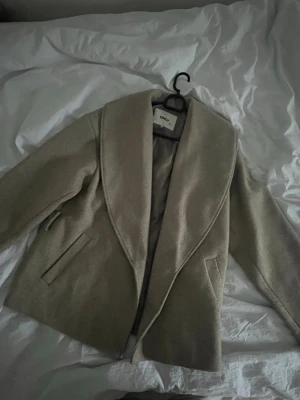 Beige kappa från ONLY LIFE(inget snöre ) - Stilren beige kappa från ONLY LIFE med bred krage och två fickor framtill. Klassisk rak passform och mjukt material som känns lyxigt. Perfekt för dig som vill ha en enkel men snygg look till hösten eller vintern.