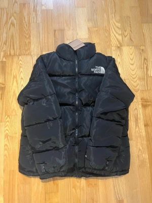 Svart pufferjacka - Säljer en svart pufferjacka med hög krage och tydlig logga på bröstet. Jackan har quiltad design, dragkedja framtill och långa ärmar. Perfekt för kalla dagar och riktigt snygg till streetwear-stilen. LITEN I STORLEKEN!