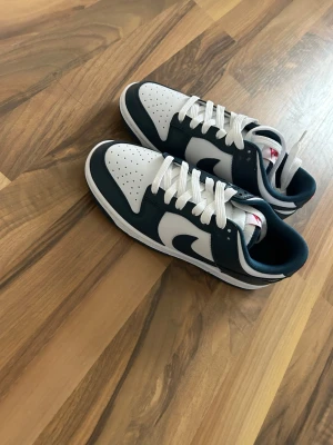 Nike Dunk Low vit/marinblå - Nike Dunk Low sneakers i vit och marinblå färg med klassisk snörning och rund tå. Skorna har en vit bas med marinblå detaljer och röd Nike-logga på hälen. Tillverkade i syntet och skinn för en clean och sportig look. Som jag fick av en kompis helt oanvända men var för små 