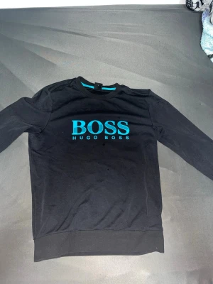 Svart sweatshirt från Hugo Boss - Svart sweatshirt från Hugo Boss med turkost tryck på bröstet där det står 'BOSS HUGO BOSS'. Tröjan har rund halsringning och ribbade muddar vid ärmslut och nederkant. Perfekt för en clean och stilren look.