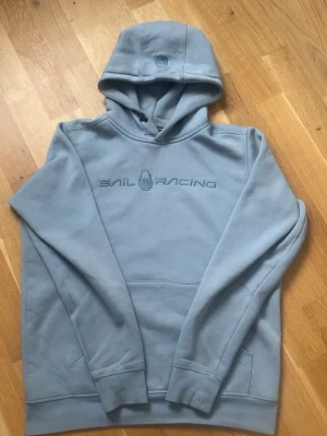 Ljusblå hoodie från Sail Racing - Ljusblå hoodie från Sail Racing med broderad logga på bröstet. Klassisk känguruficka framtill och huva med diskret broderad detalj. Tillverkad i mjukt material som känns skönt mot huden. Perfekt för en avslappnad och sportig stil. Storlek M, Vid funderingar skriv, Tack 🤗