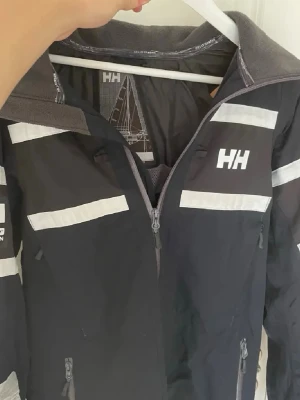 vind/seglarjacka från Helly Hansen - Snygg vindjacka från Helly Hansen med vita reflexdetaljer och dragkedja framtill. Jackan har hög krage, praktiska fickor och HH-logga på bröstet. Perfekt för blåsiga dagar och outdoor-aktiviteter. Tveka inte att skriva. Fraktar samma dag.