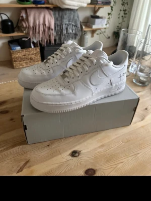 Vita Nike Air Force 1 sneakers - Klassiska vita Nike Air Force 1 sneakers i skinn med snörning och rund tå. Sidan har en unik svart textdetalj som gör dem extra speciella. Perfekta för dig som gillar stilrena och tidlösa sneakers med en twist.