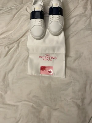 Valentino Garavani vita sneakers - Snygga vita sneakers från Valentino Garavani med bred mörkblå rem över ovandelen och ikoniska nitar på hälen. Skorna har rund tå, snörning och är tillverkade i skinn. Perfekta för dig som vill ha en clean men lyxig look.
