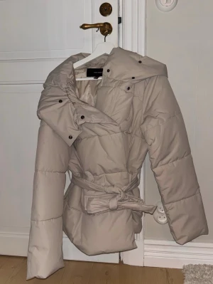 Beige pufferjacka med bälte från vero moda - Snygg beige pufferjacka från vero moda med stor krage och bälte i midjan för en trendig siluett. Jackan har quiltad design och långa ärmar, perfekt för kyligare dagar. Stängs med tryckknappar och har en oversized känsla. Köpte för 800 aldrig anbänts