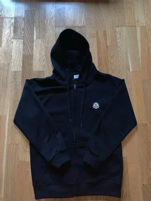 Svart zip hoodie från Moncler - Säljer en svart hoodie från Moncler med dragkedja framtill och klassisk Moncler-logga på bröstet. Hoodien har justerbar huva med snören och ribbade muddar vid ärmar och nederkant. Tillverkad i mjuk bomull för skön komfort. Vid frågor är det bara att höra av sig!
