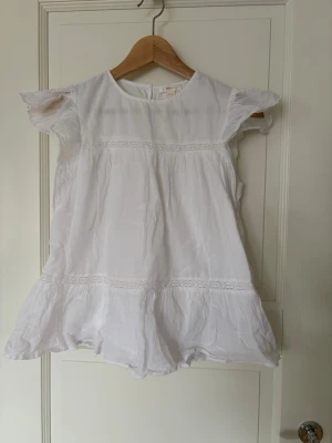 Vit volangblus från Gina Tricot - Söt vit blus från Name It med volangdetaljer och korta ärmar. Blusen har rund halsringning och fina spetsband som dekoration. Tillverkad i ett lätt och luftigt tyg, perfekt för varma dagar.