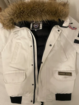 Vit dunjacka från Canada Goose - Snygg vit dunjacka från Canada Goose med svart foder och stor huva med fuskpäls. Jackan har flera fickor, dragkedja och ribbade muddar. Perfekt för kalla vinterdagar och har klassisk patch på ärmen.