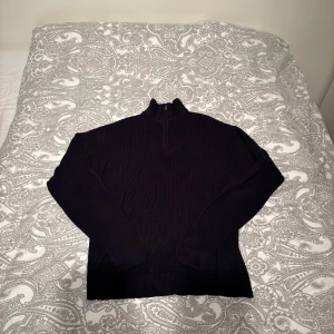 Marinblå stickad tröja med halvzip lindex  - Mysig marinblå stickad tröja med hög krage och halvzip framtill. Tröjan har lång ärm och ribbstickade muddar vid ärmslut och nederkant. Perfekt för lager på lager och en clean look.