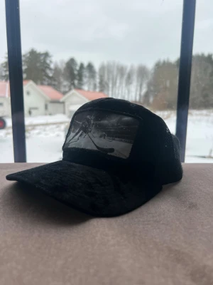 Svart truckerkeps med hockeymotiv - Säljer en svart truckerkeps med mesh på sidorna och ett stort svartvitt tryck av en hockeyspelare på framsidan. Kepsen har böjd skärm och justerbar snapback baktill. Perfekt för dig som gillar sportig stil och hockey.