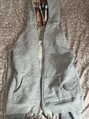 Grå hoodie från Burberry - Snygg grå hoodie från Burberry med dragkedja, två stora fickor framtill och klassiskt rutigt foder i huvan. Broderad Burberry-logga på bröstet och ribbade muddar vid ärmslut och nederkant. Perfekt för en avslappnad och stilren look.