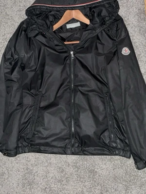 Svart vindjacka från Moncler - Snygg svart vindjacka från Moncler med huva och dragkedja framtill. Jackan har Moncler-logga på ärmen, två fickor med dragkedja och elastiska muddar. Perfekt för dig som vill ha en stilren och funktionell jacka till vardags.