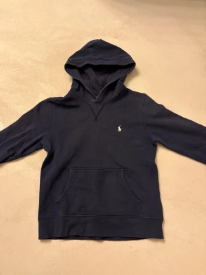 Mörkblå hoodie från Polo Ralph Lauren - Snygg mörkblå hoodie från Polo Ralph Lauren med klassisk huva och känguruficka. Liten vit logga broderad på bröstet. Tillverkad i mjukt bomullsmaterial som känns skönt mot huden. Perfekt för en avslappnad och stilren look.