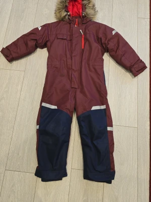 Vinröd täckbyxa med huva och reflex - Varm och tjock overall i vinrött och marinblått, perfekt för kalla vinterdagar. I mycket gott skick, endast en mycket liten repa på baksidan (syns på bilden). Använd sparsamt. Bra pris.