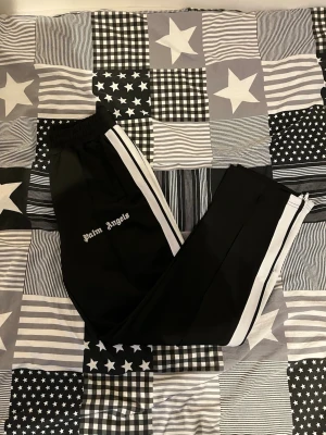 Palm Angels svarta mjukisbyxor med vita ränder - Svarta mjukisbyxor från Palm Angels med vita sidoränder och logga på vänster ben. Byxorna har elastisk midja och raka ben för en avslappnad look. Perfekta för en streetwear-stil och riktigt sköna att ha på sig.