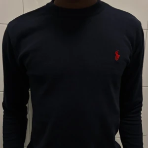 Mörkblå stickad tröja från Polo Ralph Lauren - Säljer en mörkblå stickad tröja från Polo Ralph Lauren med klassisk röd logga broderad på bröstet. Tröjan har rund halsringning, långa ärmar och ribbade muddar. Perfekt för dig som gillar stilrena och tidlösa plagg.