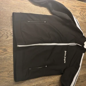Palm Angels zip tröja - Svart track jacket från Palm Angels med vita ränder längs ärmarna och vit dragkedja framtill. Jackan har ståkrage, två fickor med dragkedja och logga på bröstet. Perfekt för en sportig och trendig look. Saknar en dragkedja, Storlek Large men sitter som medium