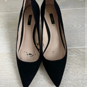 Svarta mockapumps från Mango - Stilrena svarta pumps från Mango i mocka med spetsig tå och låg klack. Klassisk och elegant design som passar till många olika outfits. Skorna har en smal siluett och är helsvarta både på utsidan och klacken. Använt skorna en gång men var aldeles för små, ser rätt slitna ut inget men ser när skorna är på