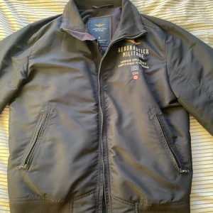 Svart bomberjacka Aeronautica Militare - Snygg svart bomberjacka från Aeronautica Militare med broderad logga och text på bröstet. Jackan har dragkedja framtill, två snedställda fickor med dragkedja och ribbade muddar vid ärmslut och midja. Perfekt för dig som gillar en clean och sportig look.