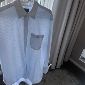 Randig skjorta från Henri Lloyd - Snygg blå- och vitrandig skjorta från Henri Lloyd med klassisk krage, bröstficka med broderad logga och kontrastdetaljer på krage och axlar. Skjortan har långa ärmar och knäppning framtill. Perfekt för en clean och stilren look.