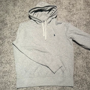 Ralph Lauren hoodie  - Snygg ljusgrå hoodie från Polo Ralph Lauren med klassisk broderad logga på bröstet. Tröjan har huva med vita snören, känguruficka och långa ärmar. Perfekt för chill dagar eller när du vill ha en clean och enkel look.