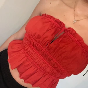 Röd volang tubtopp med snörning - Säljer en röd tubtopp med volangkant och smockade detaljer. Toppen har snörning framtill och är axelbandslös, vilket ger en snygg och trendig look. Materialet känns lätt och passar perfekt till varma dagar. Perfekt för dig som vill sticka ut med färg!