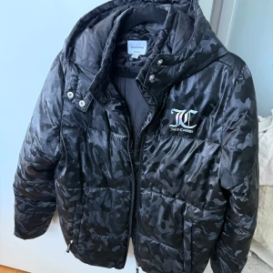 Svart camo pufferjacka Juicy Couture - Säljer en svart pufferjacka från Juicy Couture med camouflagemönster och glansig finish. Jackan har huva, dragkedja framtill och JC-logga på bröstet. Perfekt för kalla dagar och riktigt snygg streetstil.