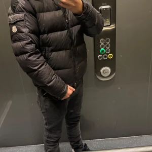 Svart Moncler pufferjacka herr - Säljer en svart pufferjacka från Moncler med klassisk logga på ärmen och dragkedja framtill. Jackan har en glansig finish, huva och quiltad design som håller dig varm under kalla dagar. Perfekt för dig som vill ha en stilren och trendig vinterjacka.