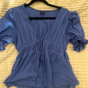 Blå blus med knyt från Gina Tricot - Supersöt blå blus från Gina Tricot med v-ringning och knyt framtill. Blusen har puffiga korta ärmar med resår och volangdetalj vid ärmslut. Tillverkad i ett lätt, strukturerat tyg. Sparsamt använd🩵