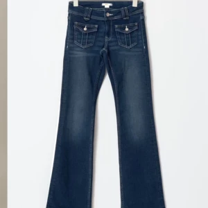 Blå bootcut jeans med fickdetaljer - Snygga blå bootcut jeans med markerade sömmar och coola fickor framtill med lock och knappar. Jeansen har klassisk femficksdesign, bälteshällor och dragkedja. Perfekta för dig som gillar en retroinspirerad look.