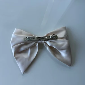 Champagnefärgat hårspänne med rosett - Snyggt hårspänne med stor rosett i champagnefärgad satin. Rosetten har en elegant glans och fästs enkelt med metallklämma.