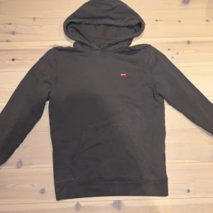 Mörkgrå hoodie från Levi's - Säljer en stilren mörkgrå hoodie från Levi's med klassisk känguruficka och liten röd logotyp på bröstet. Tröjan har huva och långa ärmar, perfekt för chill dagar. Materialet är mjukt och skönt, troligtvis bomullsmix. Storlek S.