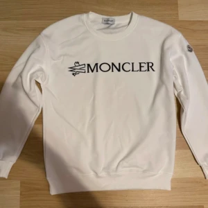 Vit Moncler sweatshirt/ M-S - Vit sweatshirt från Moncler med svart logotyp tryckt på bröstet och klassisk Moncler-patch på ärmen. Tröjan har rund halsringning, långa ärmar och ribbade muddar. Perfekt för dig som gillar stilrena och exklusiva plagg. Helt ny och oanvänd utan kvitto.